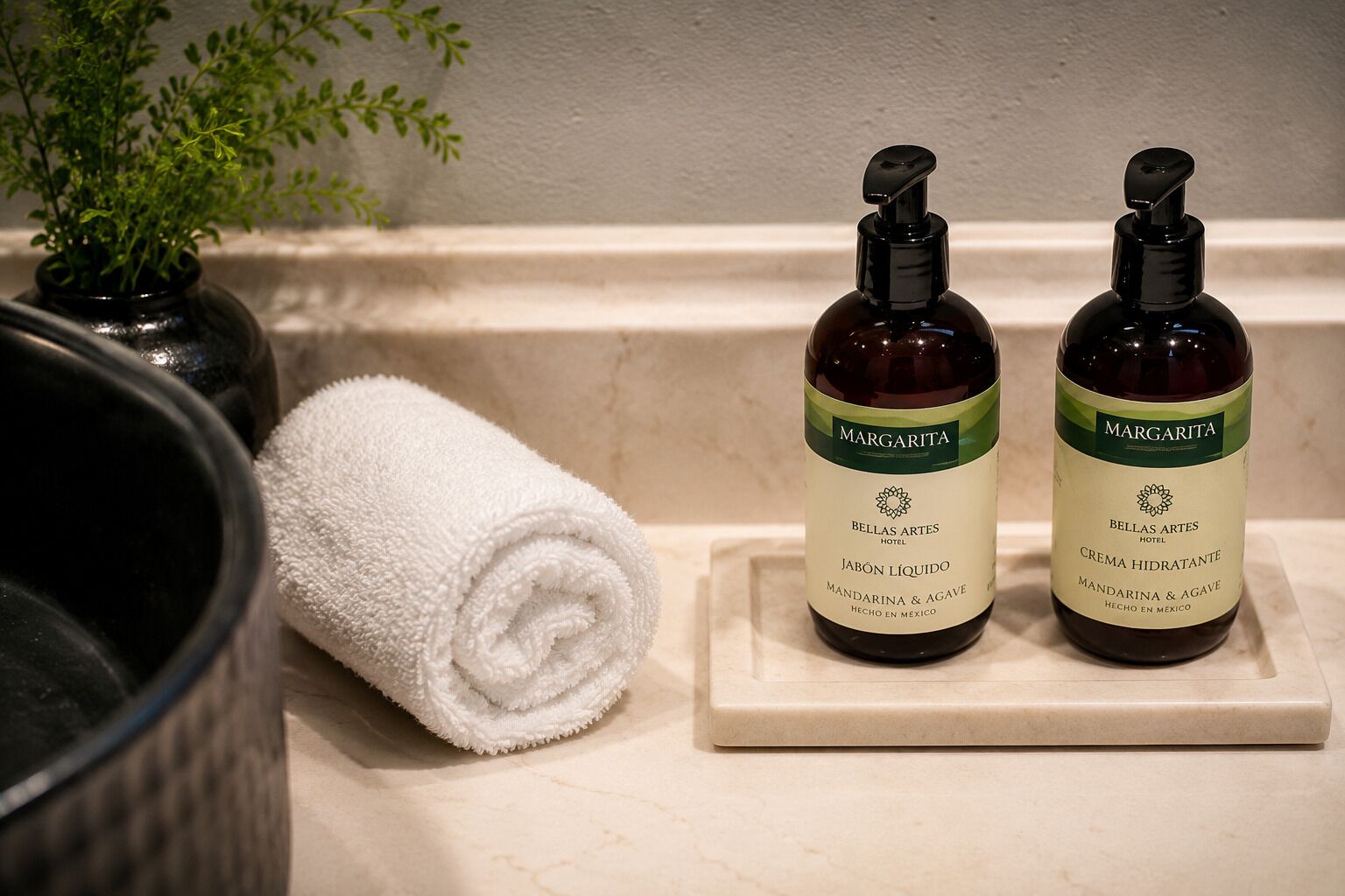 Un comptoir de salle de bain avec deux bouteilles de produits de la marque d’hôtel « Margarita », une serviette blanche roulée et une plante en pot.