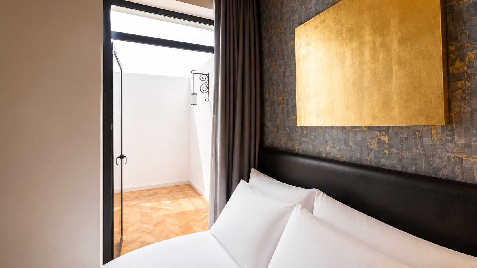 Una habitación de hotel con una cama, vista a un balcón, una lámpara fijada a la pared y una pared con textura.