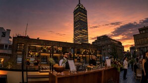 A rooftop bar with a bartender, a menu stand, and a city skyline in the background. - Hotel Bellas Artes  (Mexico City)