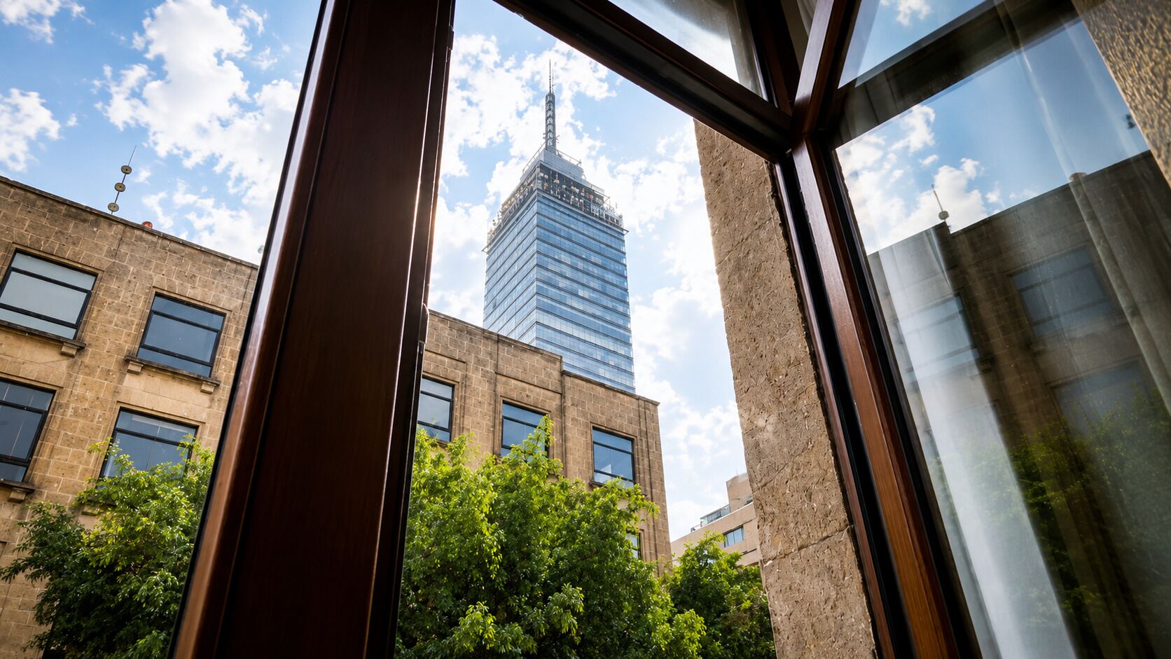 Une vue à travers la fenêtre d’un bâtiment à la façade en briques, avec un gratte-ciel en verre en arrière-plan et des arbres au premier plan.