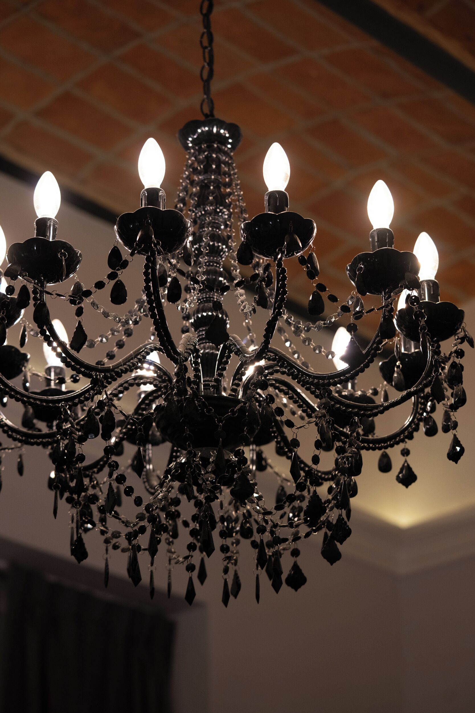 A chandelier with multiple arms and candle-like lights, hanging from a ceiling with a brick pattern.