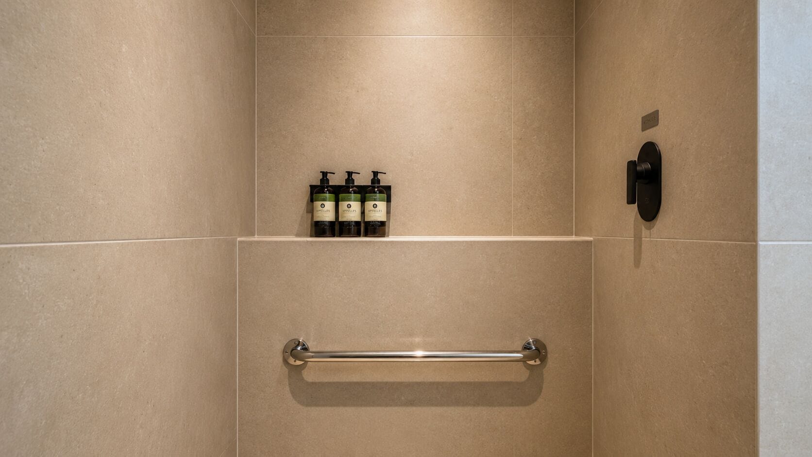 A bathroom with beige tiles, a handrail, and three bottles of toiletries on a shelf.