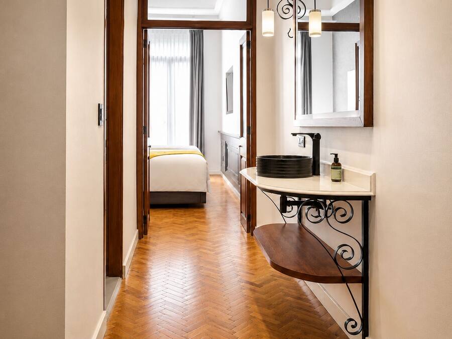 A hotel hallway with a wooden floor, a bed with a yellow blanket, a sink with a soap dispenser, and a mirror above it.