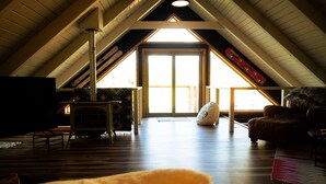 Une pièce mansardée spacieuse avec un plafond haut, des poutres apparentes, une grande fenêtre et un coin salon confortable avec un canapé et un fauteuil.