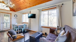 A cozy living room with a wooden ceiling, a TV mounted on the wall, a sofa with cushions, a coffee table, and a dining area with a table and chairs.