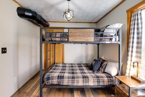 Een slaapkamer met stapelbedden, een houten vloer, een plafondventilator, een bureau met lamp en een raam met gordijnen.