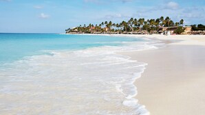 Une plage à l’eau turquoise et limpide, au sable blanc, bordée d’une rangée de palmiers et de bâtiments en arrière-plan.