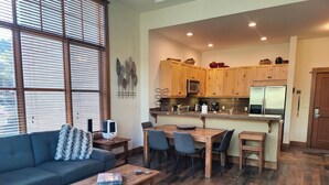 Un salon moderne avec un canapé, une table basse et un tapis. On y trouve également un coin repas avec une table et des chaises. La cuisine est équipée de meubles en bois et d’un réfrigérateur.