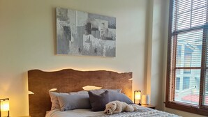 Une chambre à coucher avec une tête de lit en bois, une couvre-lit à motifs, un ours en peluche, une lampe de chevet, un tapis et une fenêtre avec des stores.