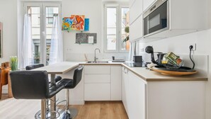 A modern kitchen with white cabinets, a built-in oven, and a dining area with a black chair and table. - Appart T2 Cosy et Chaleureux au coeur du 11eme (Paris)