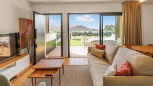 Una sala de estar moderna con sofá, mesa de centro y televisor. Cuenta con una puerta corrediza de vidrio que da a un balcón con mobiliario exterior.