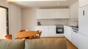 Una cocina moderna con armarios blancos, horno integrado y cafetera. Hay un comedor con una mesa redonda y sillas de madera.