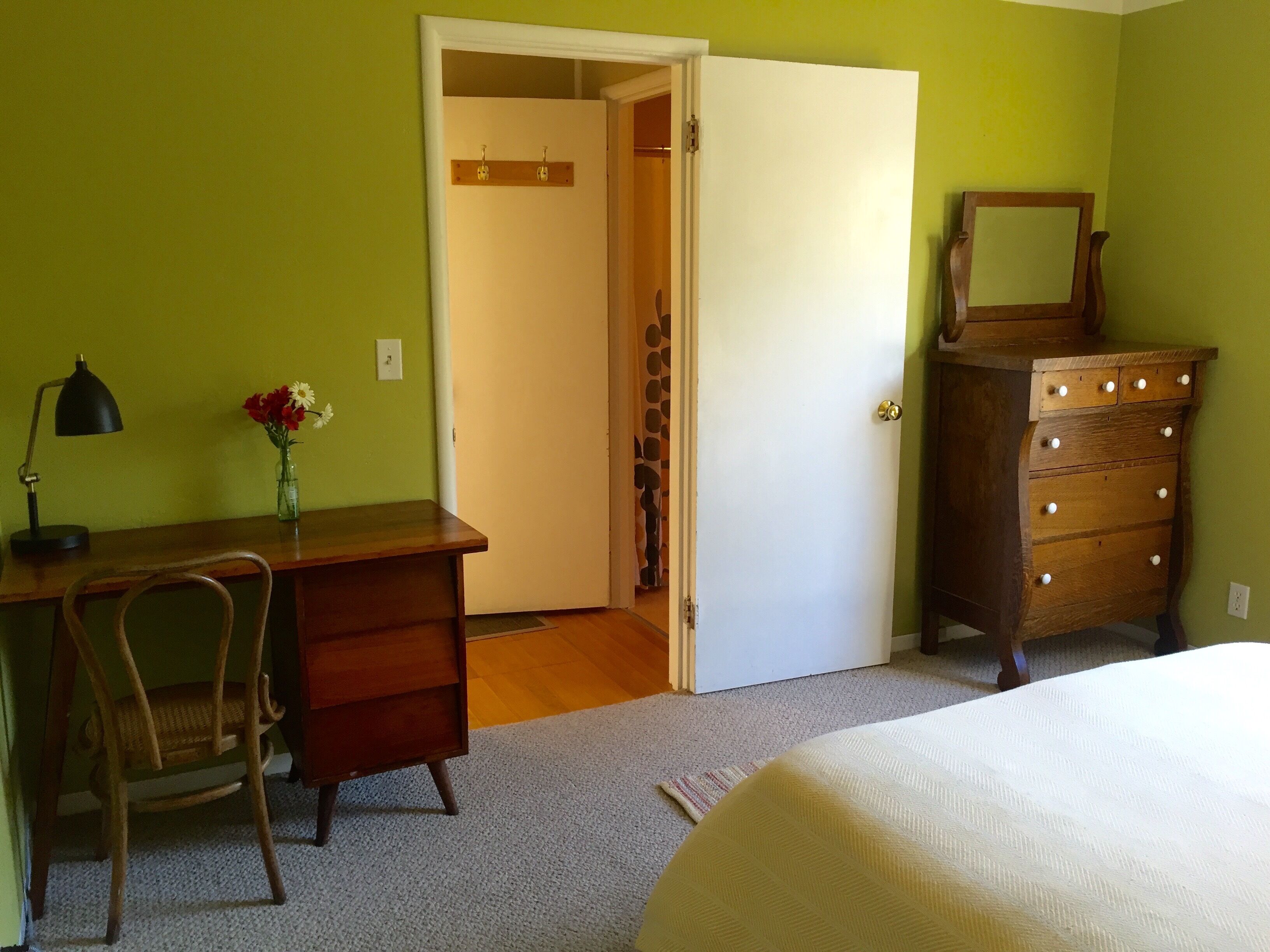 Ein Schlafzimmer mit einem Bett, einer Holzkommode, einem Schreibtisch mit Stuhl, einer Lampe und einer Vase mit Blumen.