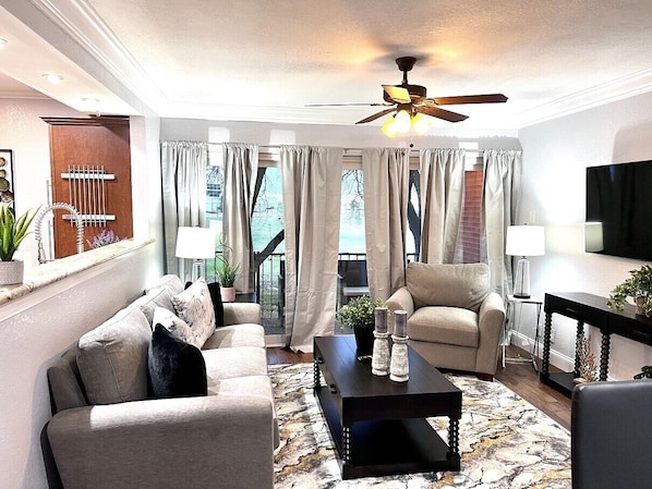 A living room with a sofa, armchair, coffee table, and a TV. There is a ceiling fan, curtains, and a plant on the windowsill.
