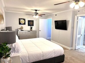 A bedroom with a bed, a ceiling fan, a TV, a dresser, and a door leading to another room.