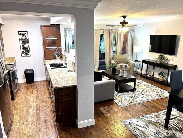 A modern kitchen with a wooden countertop, a dining area with a sofa and a coffee table, and a living room with a TV and a ceiling fan.