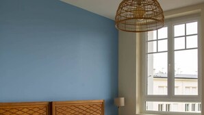 Un dormitorio con cabecera de madera, un ventanal con marcos blancos y una lámpara de techo tejida.