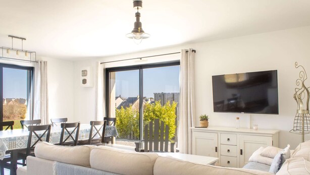 Un salón luminoso con sofá, mesa de comedor, sillas, televisor y un amplio ventanal con vistas a zonas verdes y edificios.