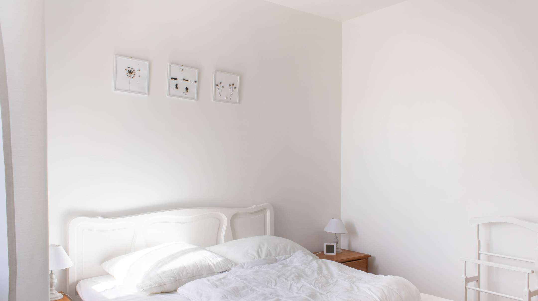 A minimalist bedroom with a white bed, a bedside table with a lamp, and three framed pictures on the wall.