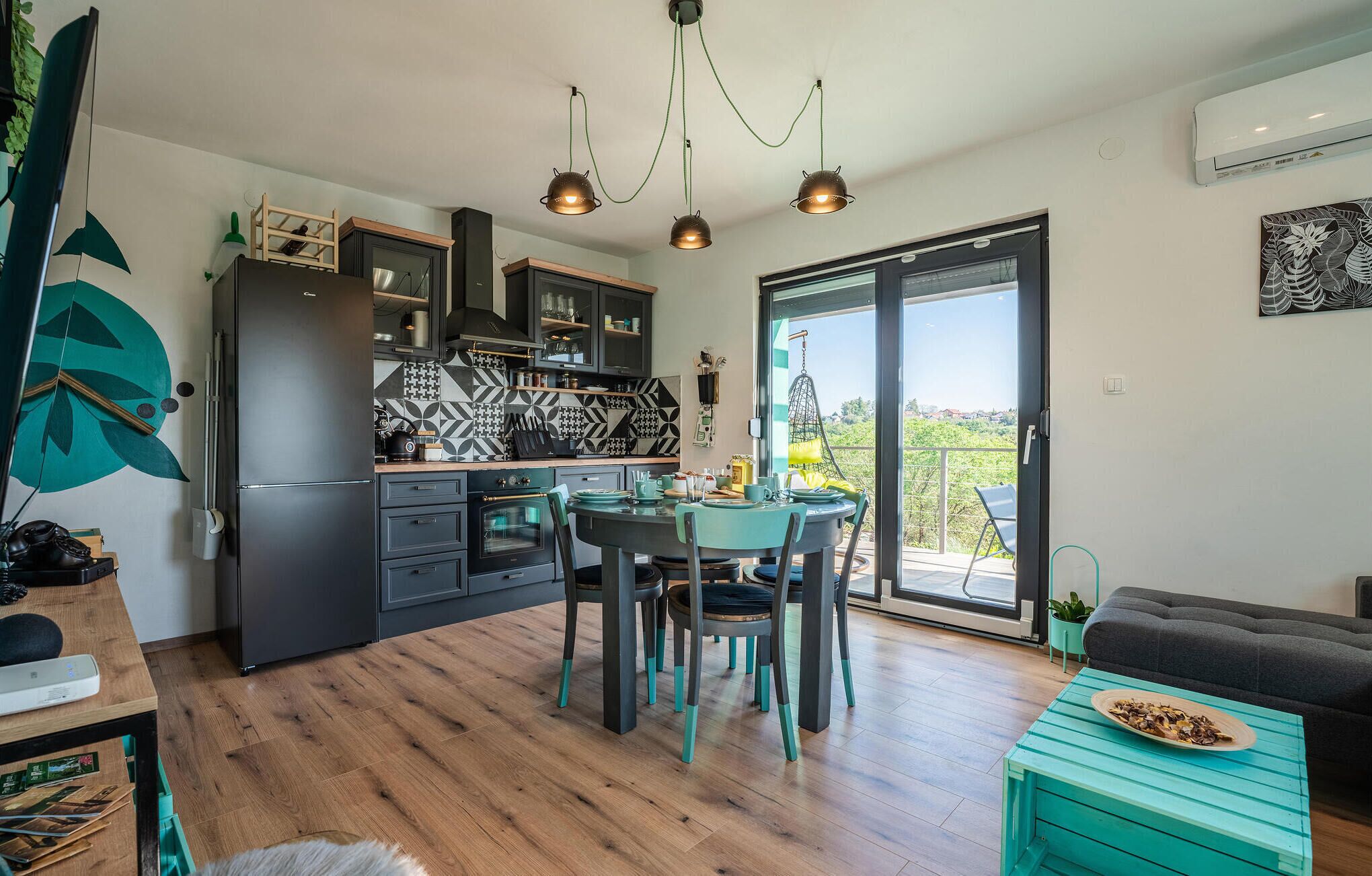A modern kitchen with dark cabinetry, a dining area with teal chairs, and a balcony with a hanging chair.