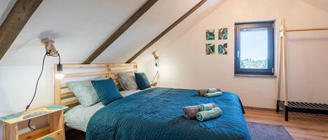Ein gemütliches Schlafzimmer im Loft-Stil mit schrägem Dach, einem Holzbettgestell, einer blauen gesteppten Bettdecke und einem kleinen Nachttisch mit Lampe.