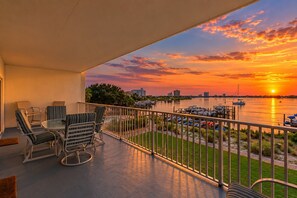 Un balcon agrémenté de mobilier d’extérieur, une vue sur un coucher de soleil se reflétant sur l’eau et un port de plaisance avec des bateaux.