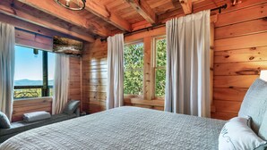 A wooden cabin bedroom with a large bed, two windows with curtains, and a view of the outdoors. - Cabin With Amazing Mountain Views! Sleeps 8! 8 Mins From The Strip! (Sevierville)