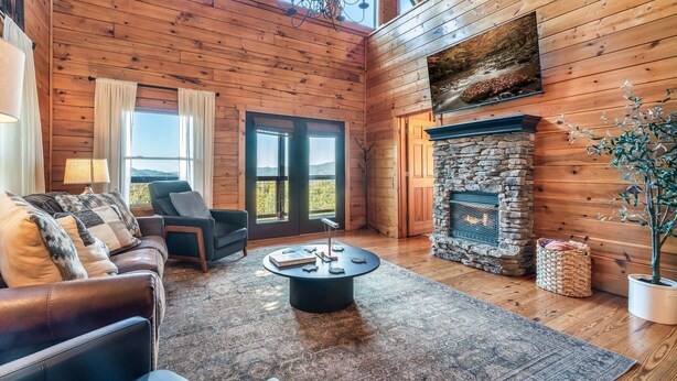 A cozy log cabin interior with a stone fireplace, leather sofa, and a large window offering a view. - Cabin With Amazing Mountain Views! Sleeps 8! 8 Mins From The Strip! (Sevierville)