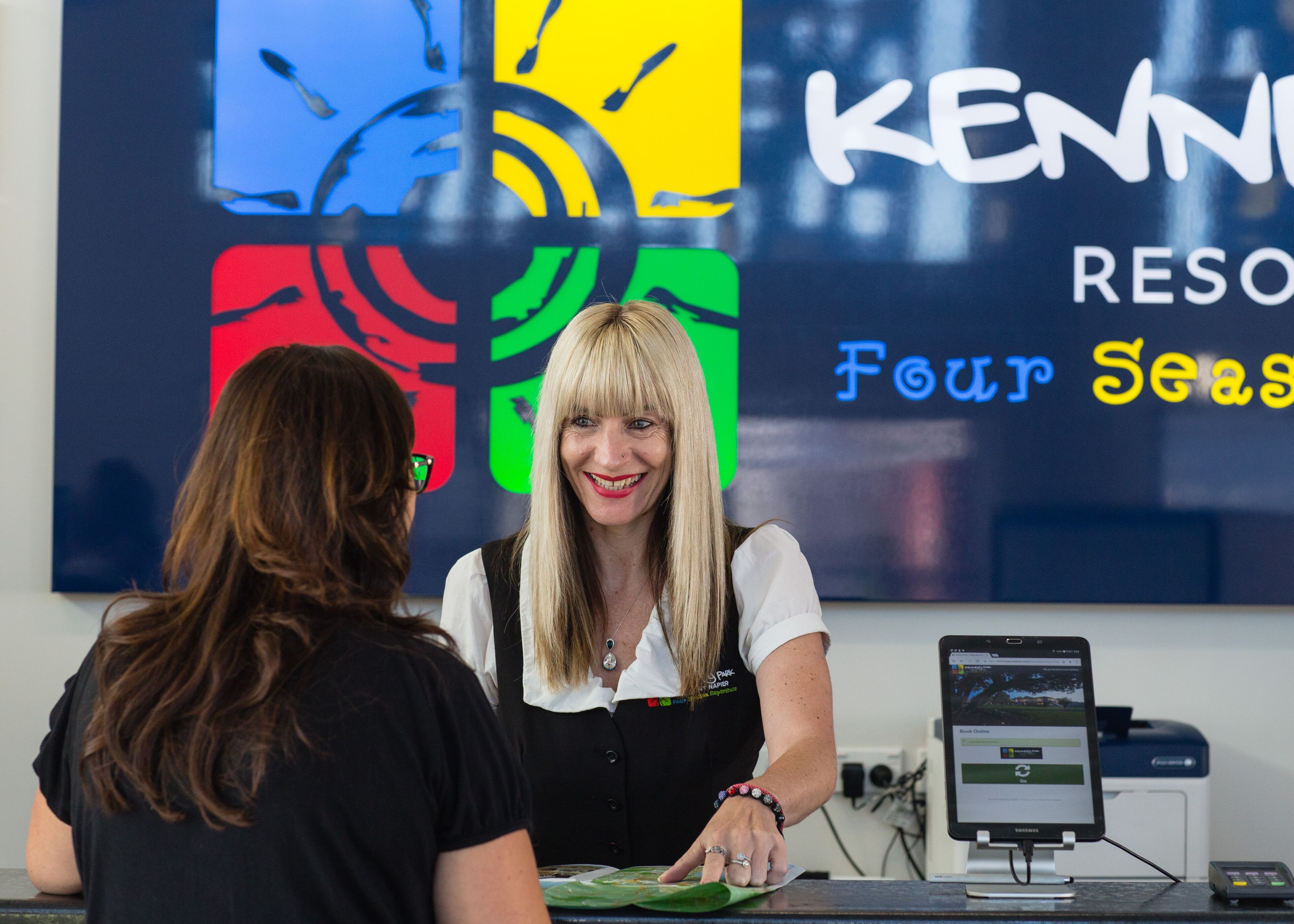 A hotel reception desk with a computer monitor, a point-of-sale terminal, and a colorful wall with the text "KENN RESORT" and "Four Seasons".
