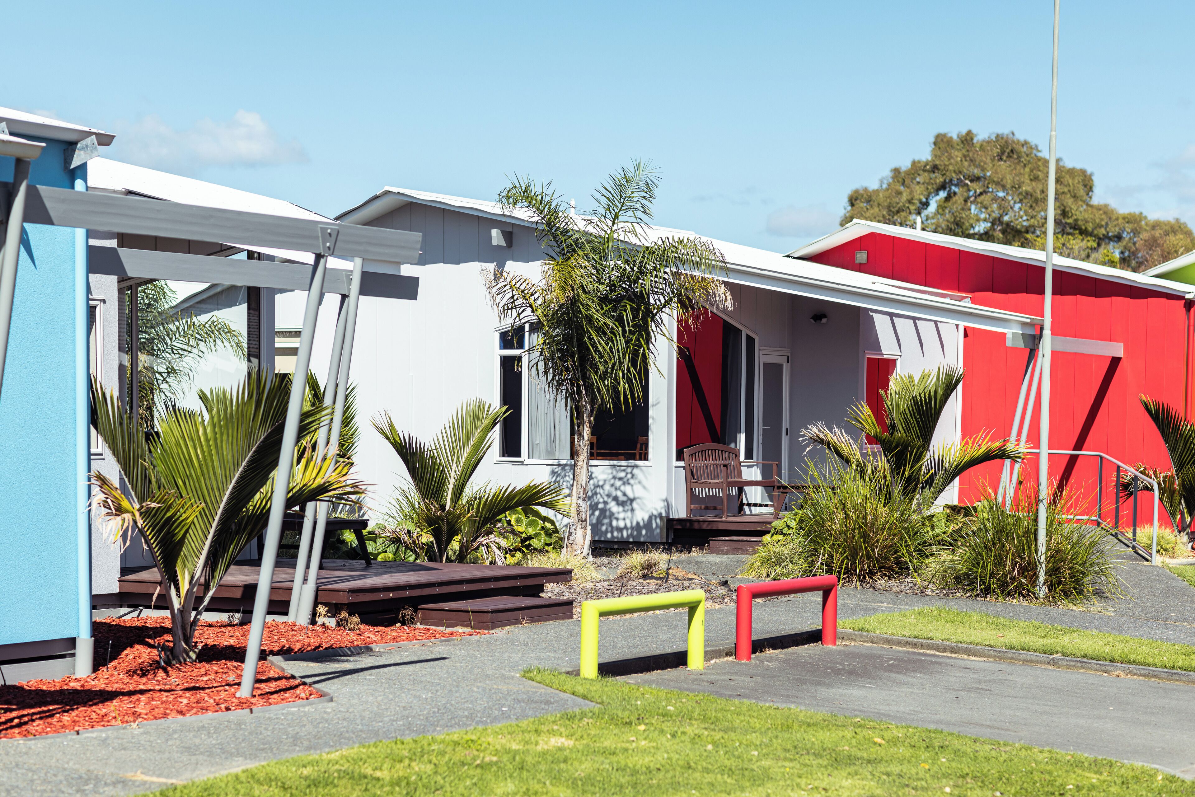 Un edificio moderno con facciata rossa e bianca, una zona esterna coperta e un giardino curato con palme.