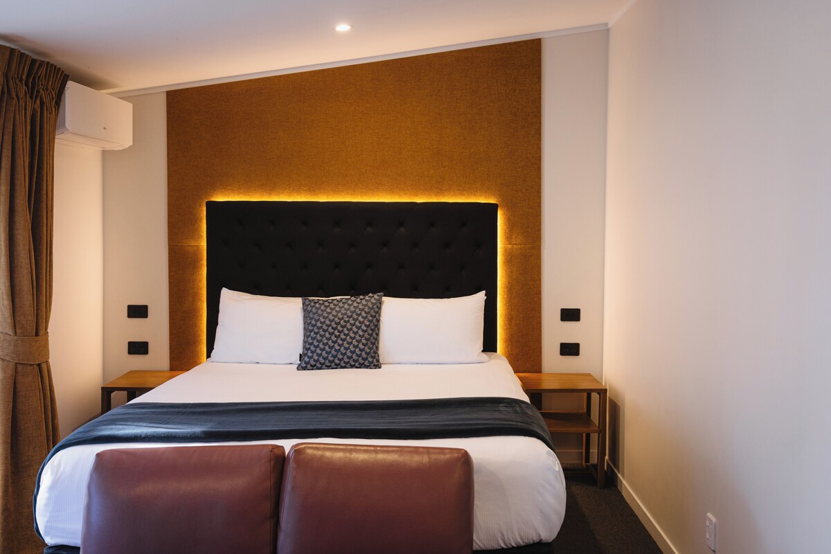 A hotel room with a large bed, a headboard with a light panel, two bedside tables, and a brown curtain.