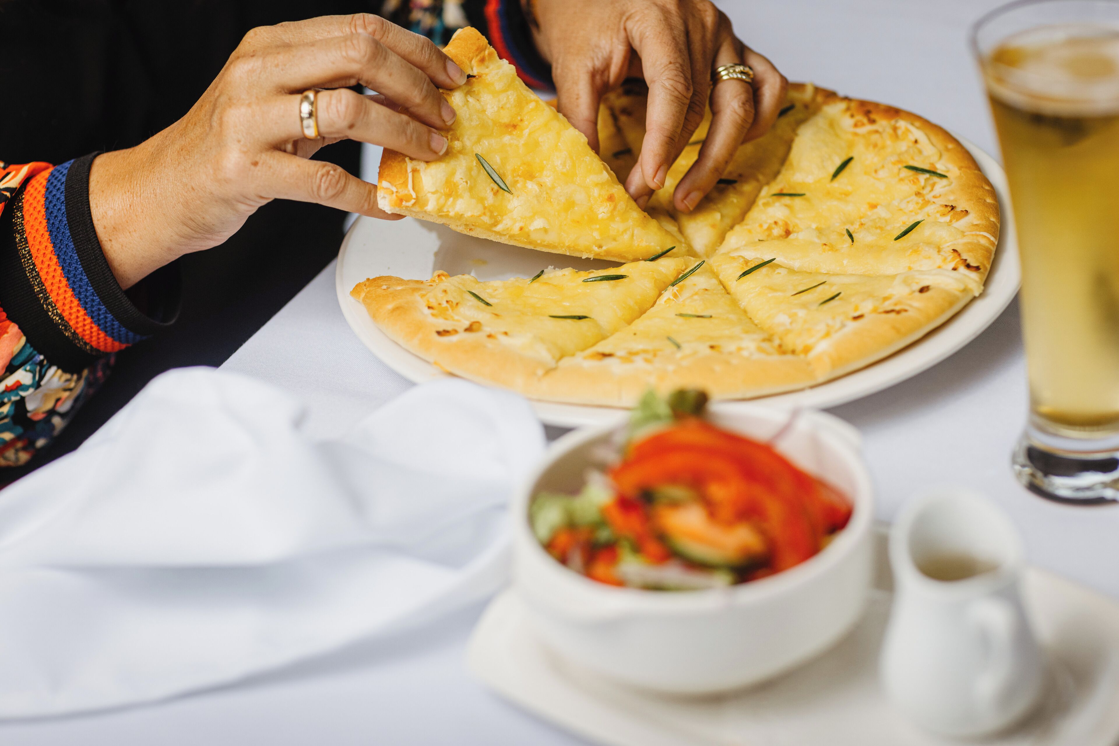 Una persona che tiene una fetta di pizza con un piatto bianco e un bicchiere di una bevanda gialla su un tavolo.