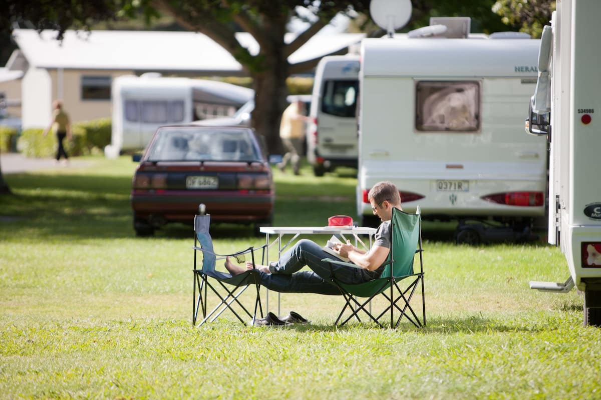 A person sitting on a lawn chair at a table with a book, surrounded by RVs and a car in a grassy area.
