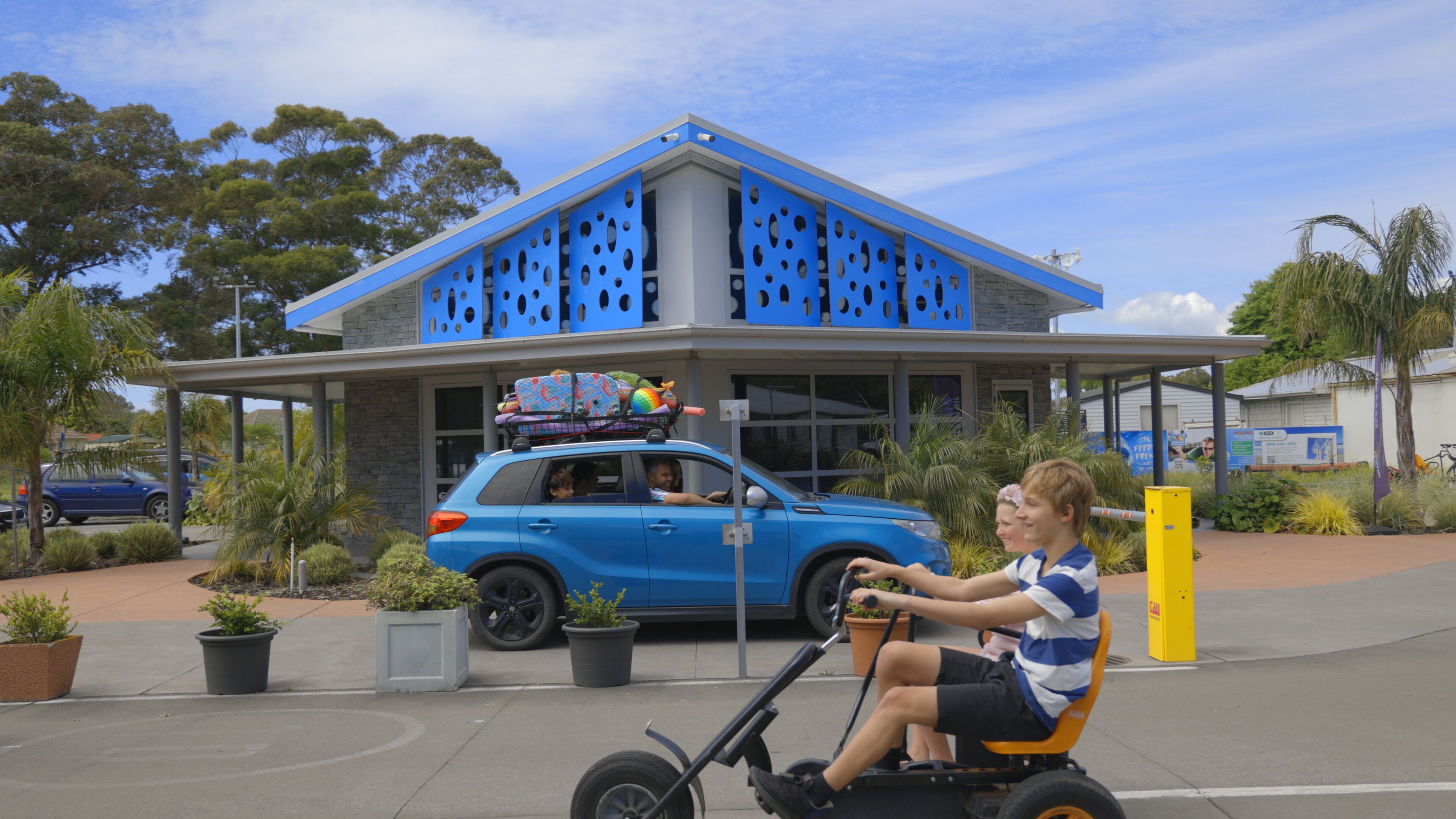 Un edificio moderno con paneles azules perforados, un coche azul con un juguete de colores en el techo, y una persona montando en un scooter.