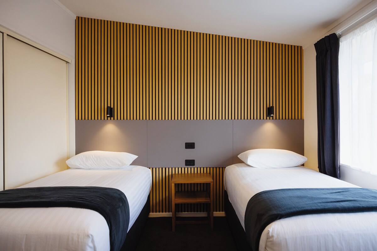 A hotel room with two single beds, a small wooden table, and a slatted wall design.