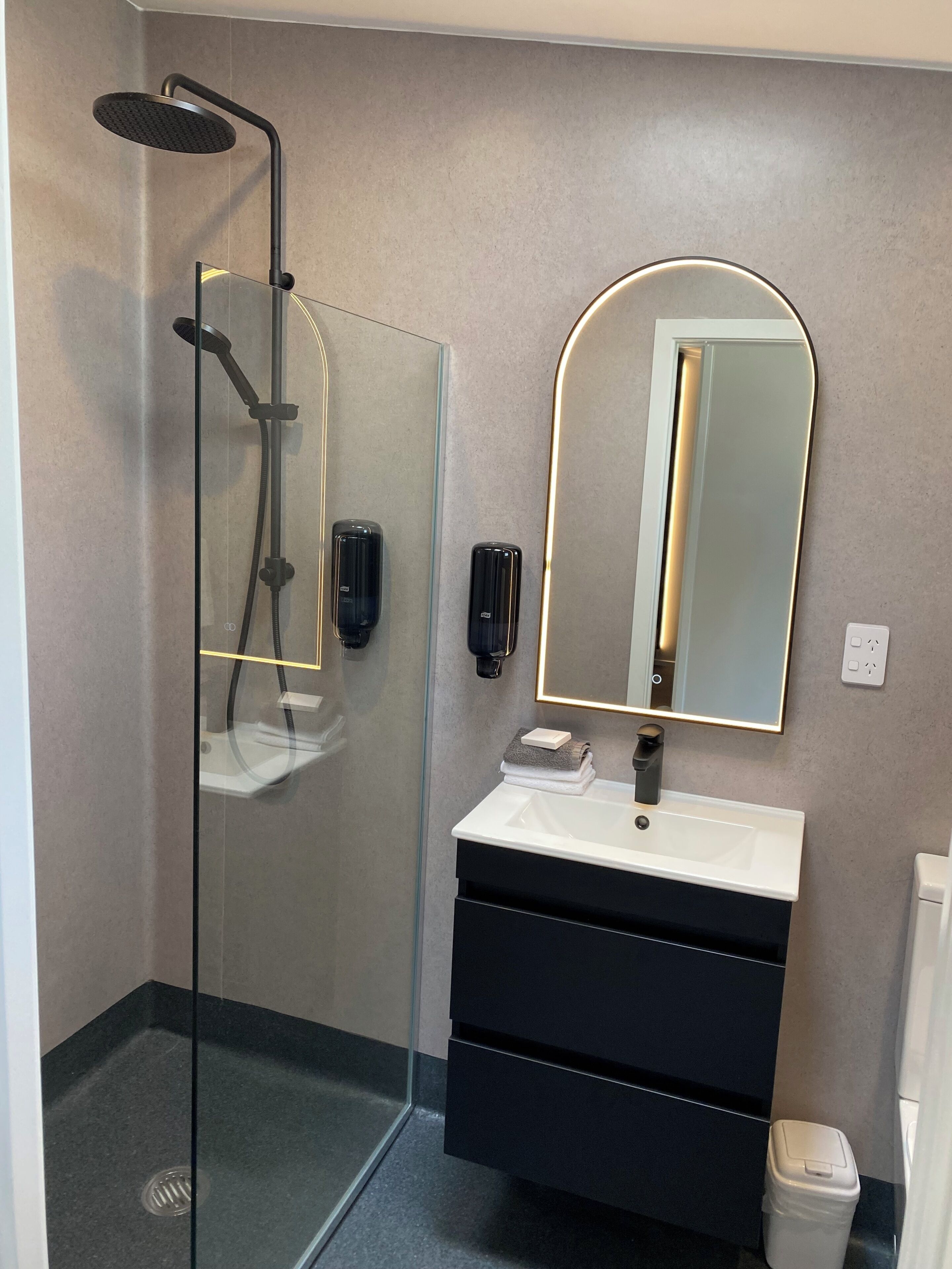 A modern bathroom with a glass-enclosed shower, a white sink with a black cabinet, a large mirror, and a wall-mounted soap dispenser.
