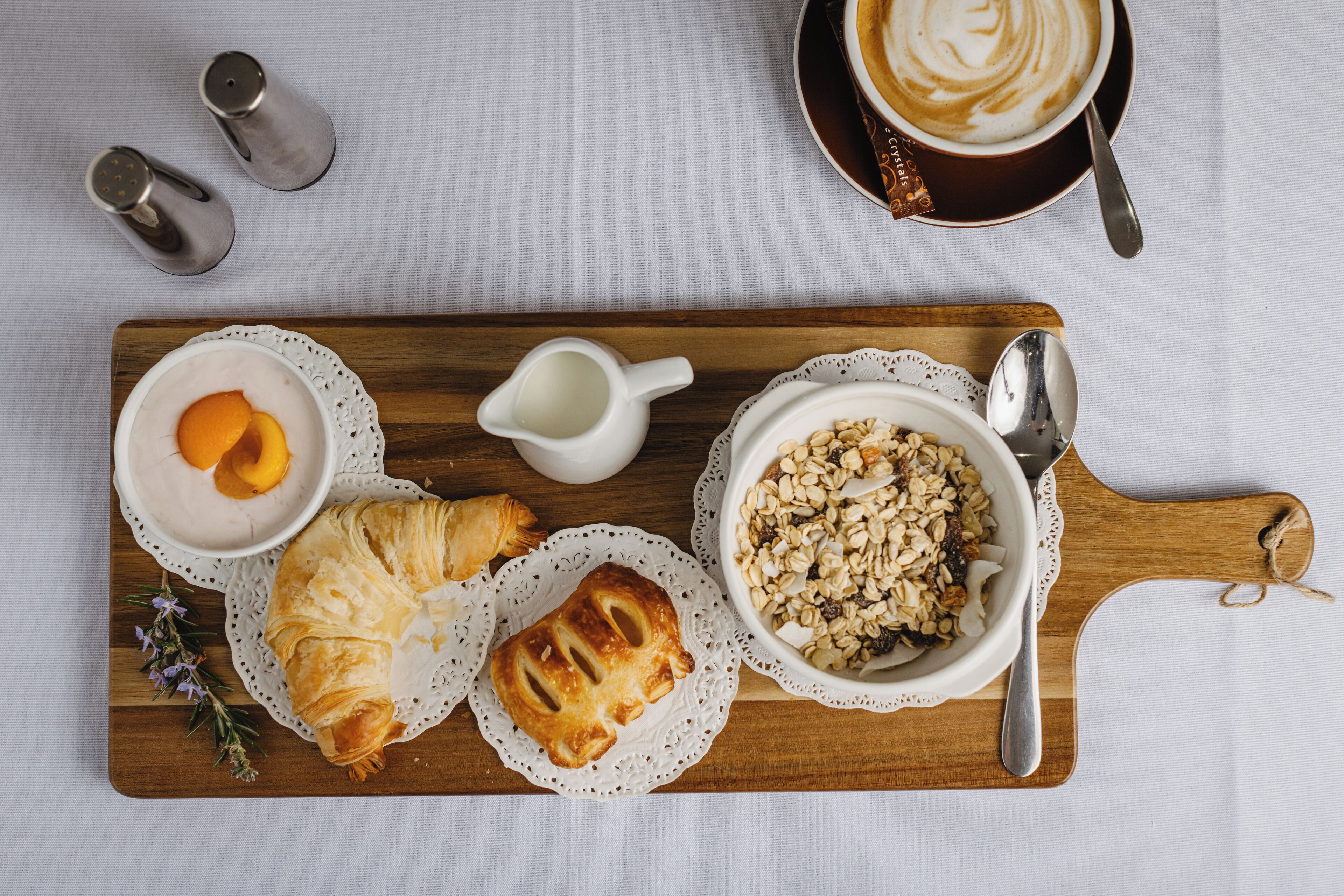 A breakfast tray with croissants, a bowl of cereal, a cup of coffee, and a small jug of milk on a wooden board.