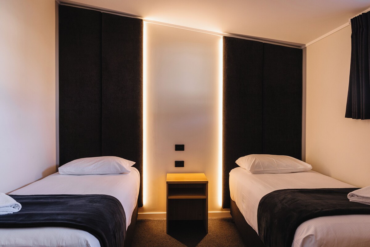 A hotel room with two beds, a nightstand, and a wall-mounted light.