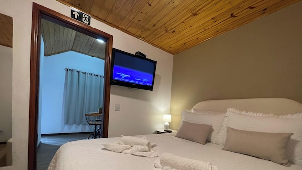 A hotel room with a large bed, a television mounted on the wall, a wooden ceiling, and a door leading to a balcony. - Pousada Flor de Lua Monte Verde (Camanducaia)