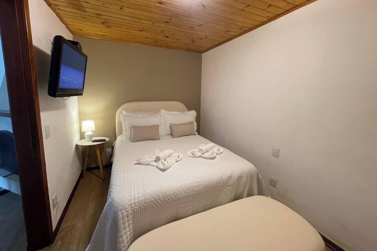 A hotel room with a wooden ceiling, a bed with white bedding and towels, a small bedside table with a lamp, and a television mounted on the wall.