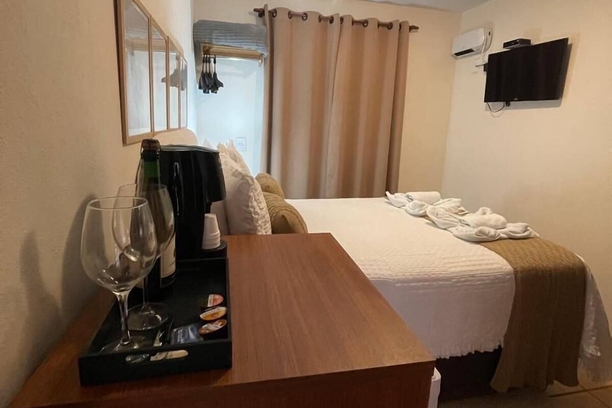 A hotel room with a bed, a wooden desk with a tray of wine glasses and snacks, a flat-screen TV mounted on the wall, and a window with curtains.