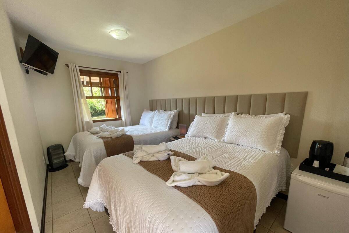 A hotel room with two beds, a TV mounted on the wall, a small refrigerator, and a window with curtains.