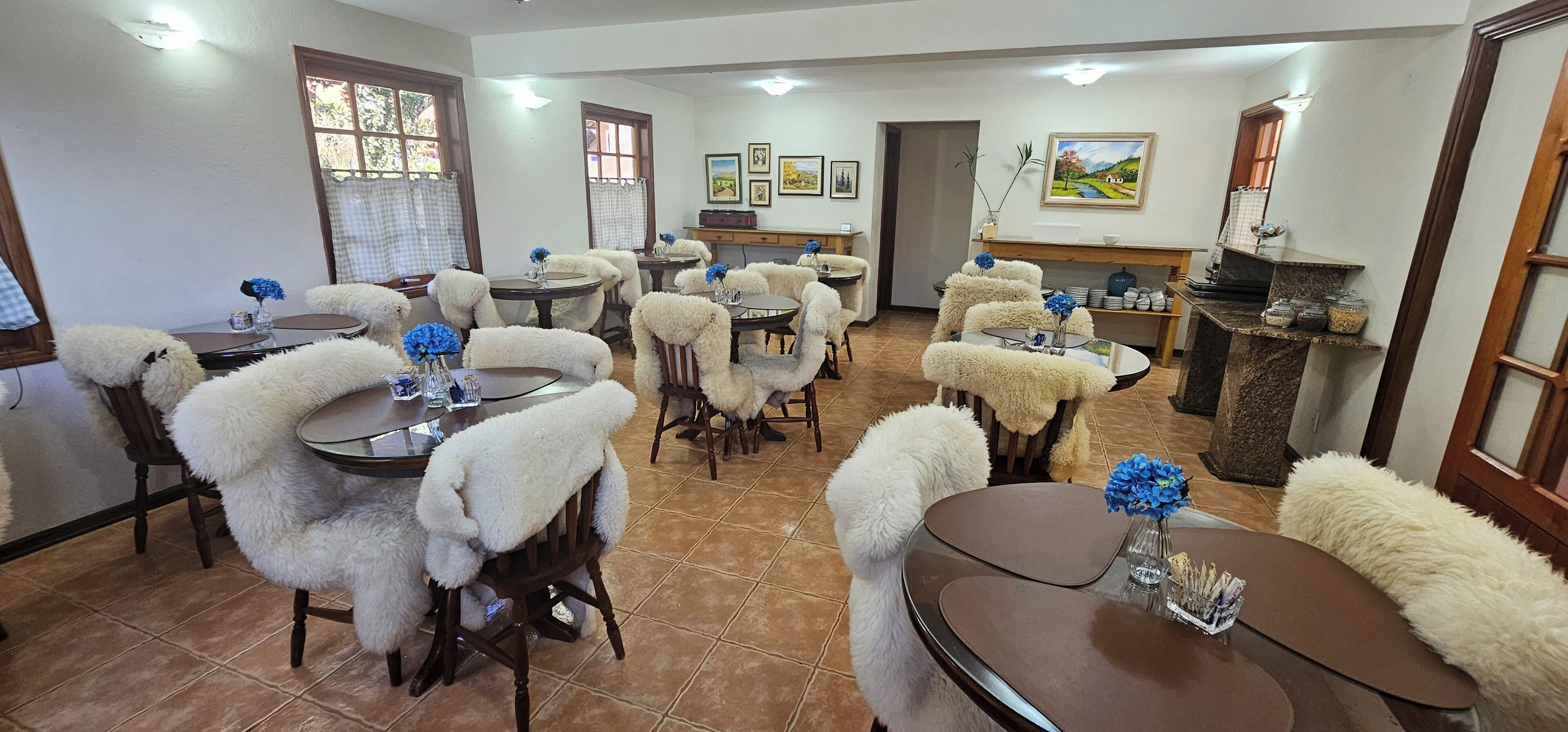 Uma área de refeições com mesas e cadeiras redondas, cada uma com uma capa branca e fofa. Há quadros nas paredes e um balcão de madeira com potes.