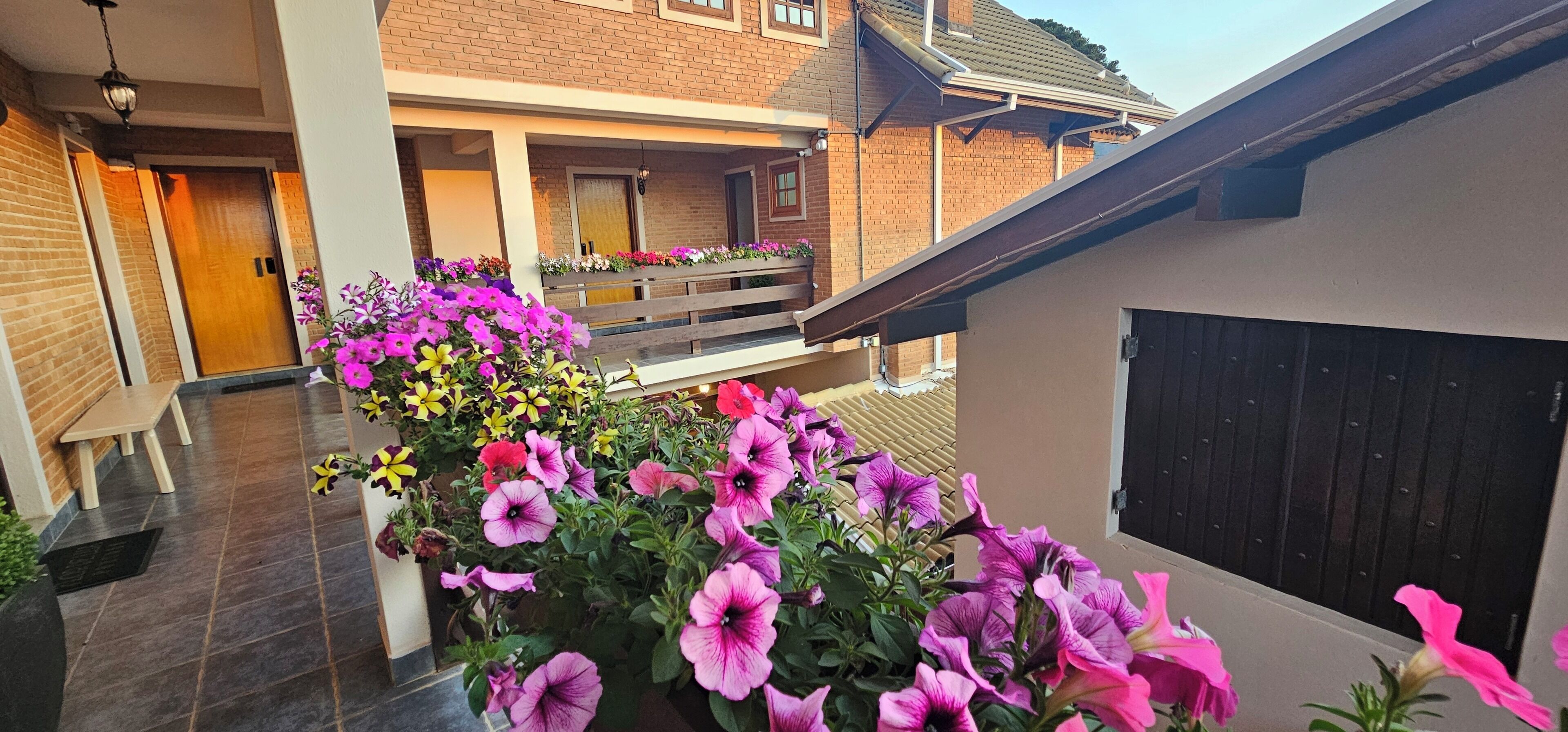 A covered outdoor area with a brick building, a wooden bench, and potted plants with pink flowers.