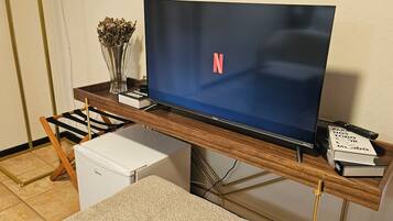 Una habitación con un mueble bajo de madera para televisor, un televisor de pantalla plana, un refrigerador blanco pequeño, un jarrón con flores secas y algunos libros.