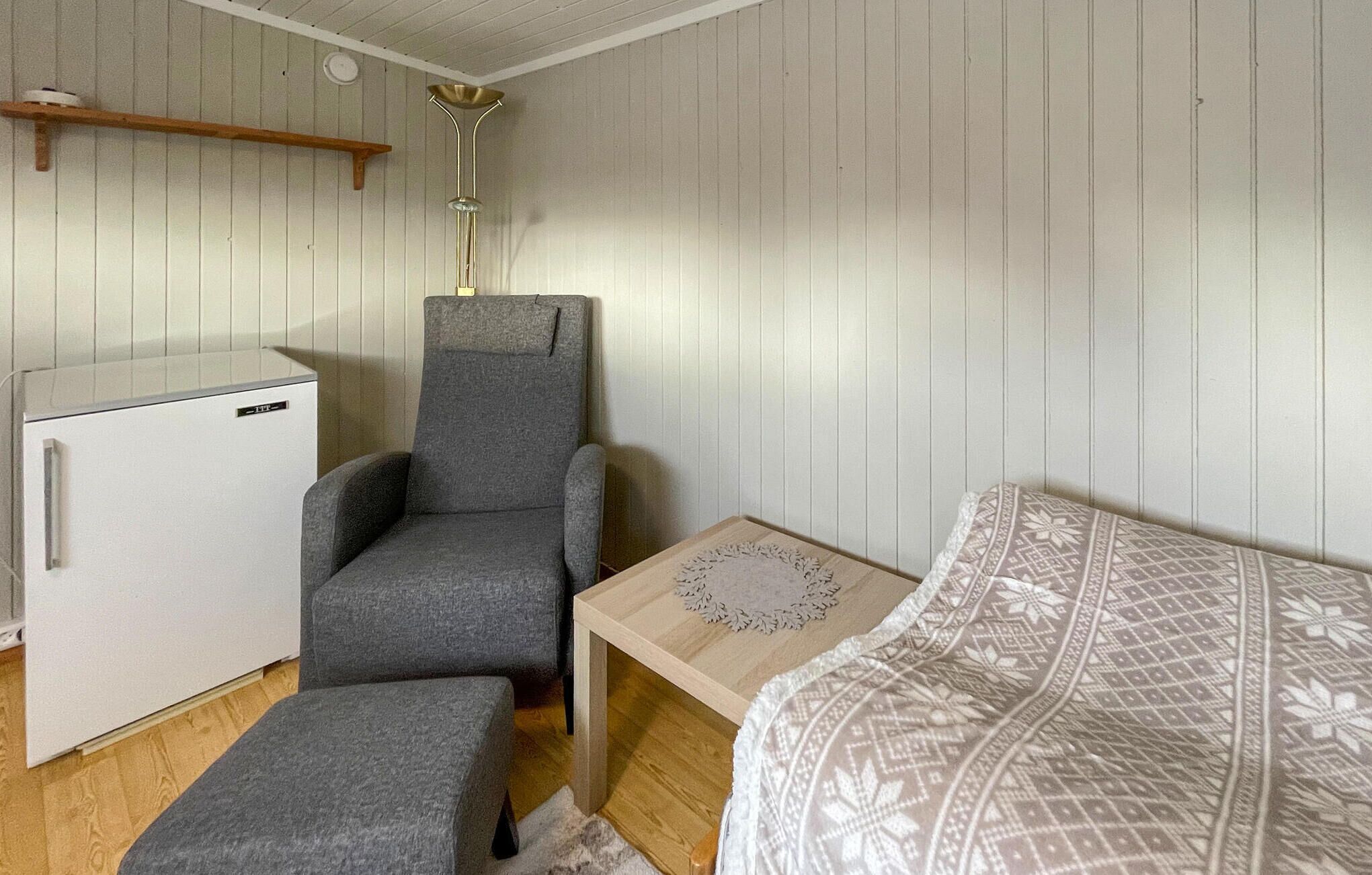 Una habitación pequeña con un sillón gris, un refrigerador blanco compacto, una mesita de madera con un mantel de encaje y una cama estampada.