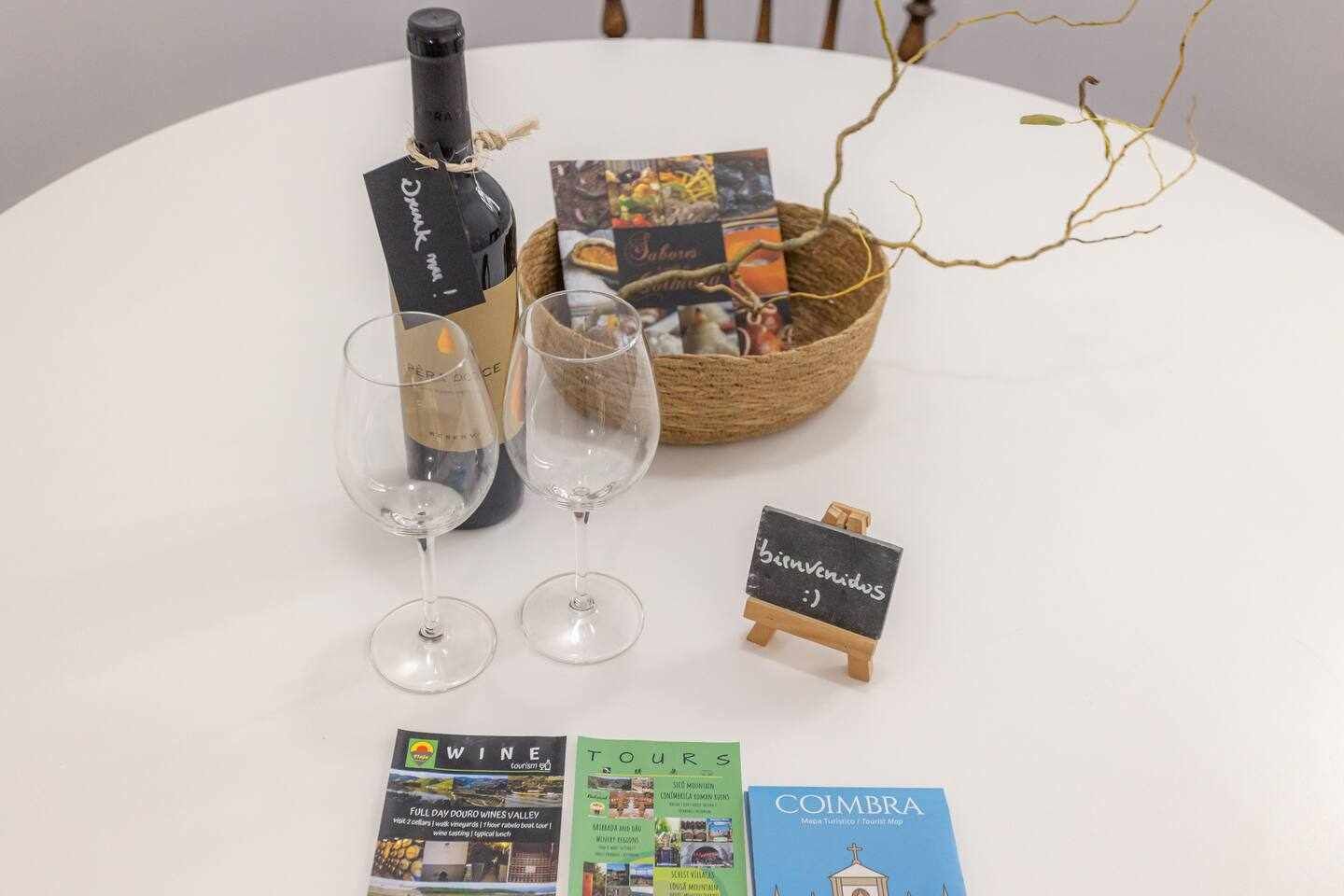 Een tafel met een fles wijn, twee glazen, een klein schoolbord waarop 'bienvenidos :)' staat, een mand met folders en een boek getiteld 'COIMBRA'.