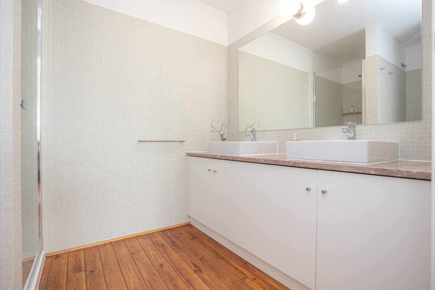 Une salle de bain avec un plancher en bois, des murs recouverts de carreaux blancs, un grand miroir et deux lavabos.