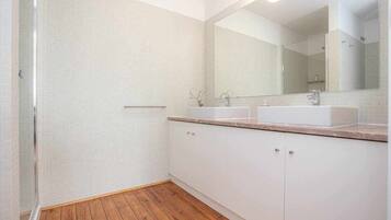 Une salle de bain avec un plancher en bois, des murs recouverts de carreaux blancs, un grand miroir et deux lavabos.