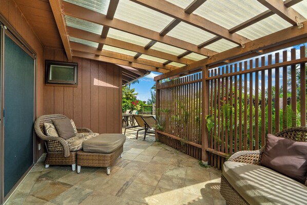 A covered patio area with wicker furniture, a television mounted on the wall, and a view of the garden.